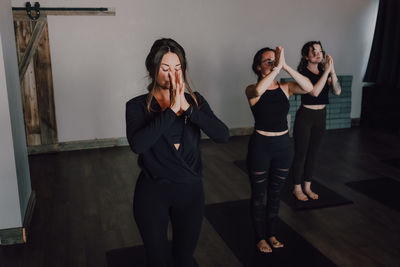 High angle of calm slender ladies in activewear with namaste standing in mountain position on sports mats on wooden floor and meditating in spacious contemporary gym with closed eyes