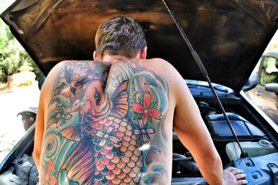 Rear view of shirtless man with tattoo on back repairing car