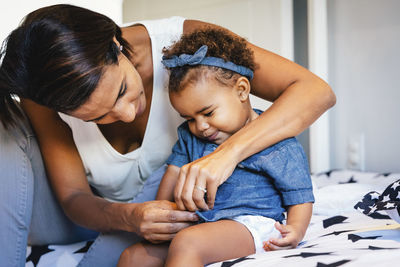 Mother dressing cute daughter while sitting on bed at home