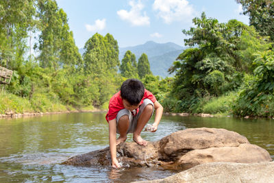 Boy crouching on rock in river against sky