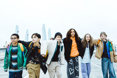 Multiracial male and female friends with arms around walking on street against clear sky