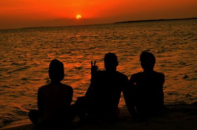 Rear view of silhouette men sitting at beach during sunset