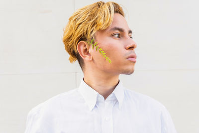 Thoughtful young male in formal shirt and small green twig on cheek looking into distance while standing on white background