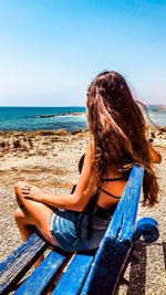 Side view of woman sitting on bench at beach against clear sky