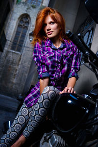 Low angle view of young woman sitting on motorcycle by building
