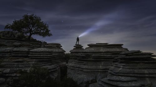 Silhouette man with illuminated flashlight standing on cliff against sky at night