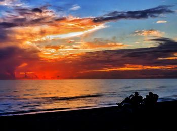 Silhouette people sitting on beach against sky during sunset