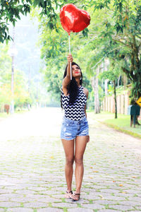 Woman holding red heart shape balloon while standing on footpath