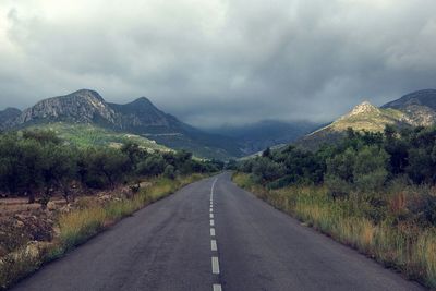 Country road leading towards mountain against cloudy sky