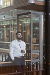 Young thoughtful businessman with headphones using smart phone while standing at office