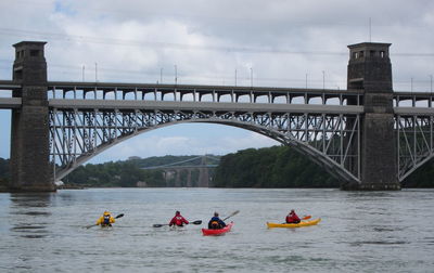 Rear view of men kayaking by britannia bridge against sky on river