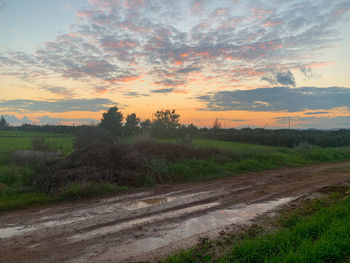 Dirt road amidst field against sky during sunset