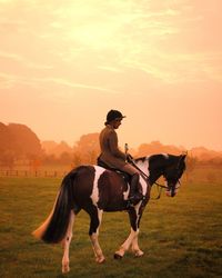 Woman riding on horse at field against sky during sunrise