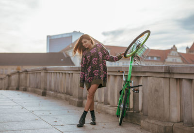 Young woman holding bicycle while standing against railing in city