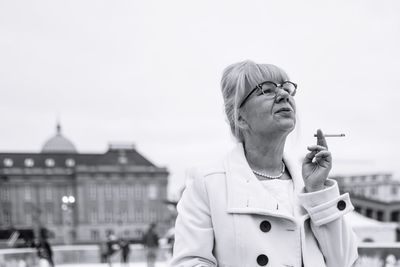 Mature woman smoking against sky in city