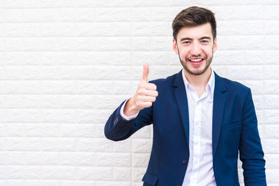 Portrait of confident man gesturing thumbs up while standing against wall