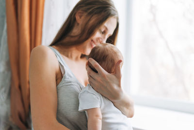 Young woman mom with long hair holding baby girl on hands near window at home