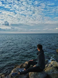 Man sitting on rock looking at sea against sky