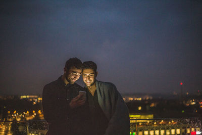 Man using smart phone while standing by male friend on terrace at night