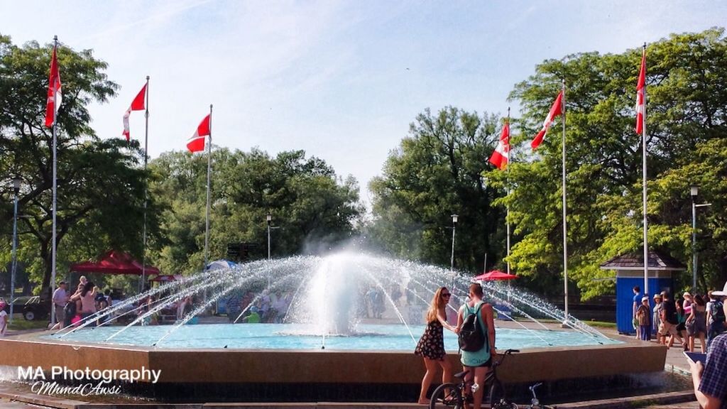 tree, lifestyles, leisure activity, water, person, men, large group of people, enjoyment, sky, mixed age range, tourism, vacations, flag, motion, railing, fun, park - man made space, fountain, tourist