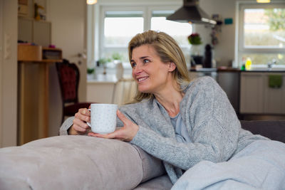 Smiling mature woman holding coffee cup while looking away on sofa at home