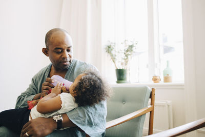 Caring father feeding milk to daughter while sitting in living room at home