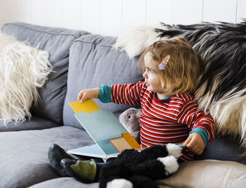 Full length of cute girl with book looking away while sitting on sofa at home