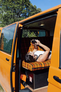 Side view of female traveler lying in van and looking through taken pictures on photo camera during summer vacation