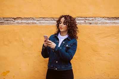 Young woman using mobile phone while standing against wall