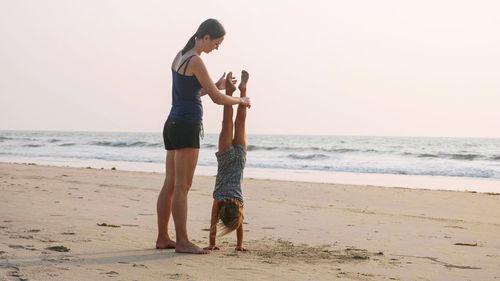 Full length of mother helping daughter in doing handstand on beach against clear sky