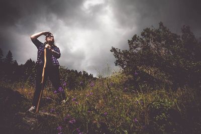 Low angle view of woman standing while shielding eyes amidst plants against sky