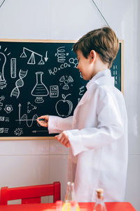 Side view of boy wearing lab coat while writing on blackboard in classroom