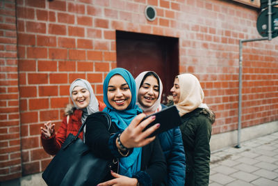 Happy young muslim woman taking selfie with female friends on sidewalk against building in city