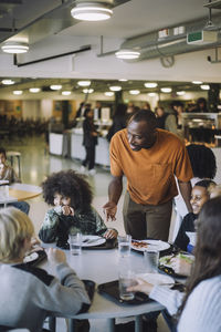 Teacher talking to students sitting at table during lunch break in school cafeteria