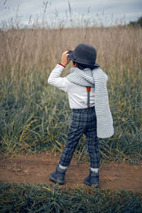 Boy child in plaid pants, hat, suspenders and scarf stands in a field in autumn rear