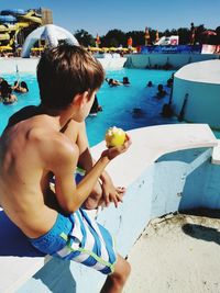 Rear view of shirtless boy eating an apple at swimming pool