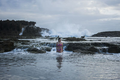 Woman stands in water near exposed rock on beach with wave crashing behind her