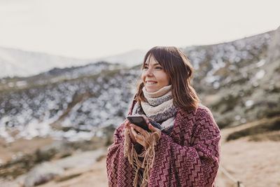 Woman looking away while holding mobile phone against mountains and sky