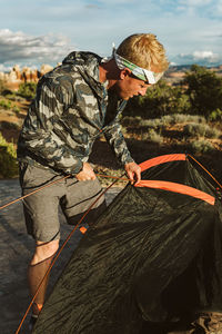 Male hiker in camo jacket and headbank sets up tent in desert