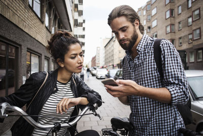 Man showing phone to female friend with bicycle at sidewalk in city