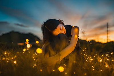 Woman with illuminated string light sitting on grass during sunset