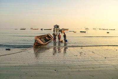 Men standing by boat on shore at beach against sky during sunset