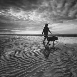 Woman walking with dog at beach against cloudy sky