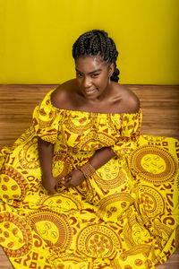 Young beautiful woman sitting on wooden floor, wearing colorful dress. against yellow background.