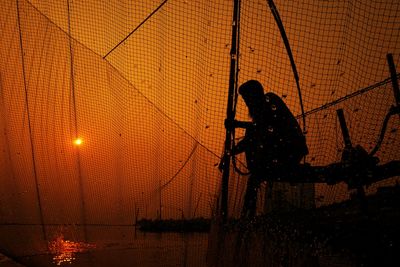 Silhouette man against orange sky seen through fence during sunset