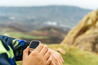 Crop of young ethnic sportswoman in activewear checking time on smart watch during outdoor training in mountainous terrain against cloudy sky