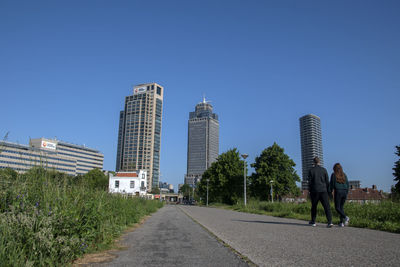 Rear view of people walking by modern buildings against clear sky