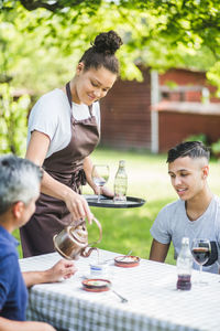 Confident young female waitress serving drink for male customers sitting at table