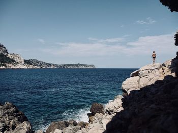 Rear view of shirtless man standing on rock formations by sea against sky