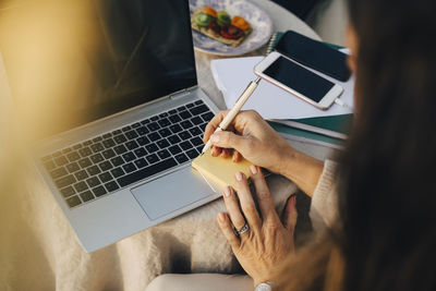 Cropped woman writing on adhesive note over laptop at table